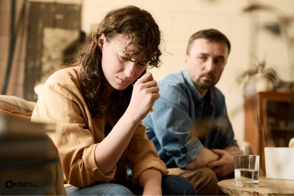A woman looks pensive while a man watches her with concern in a dimly lit room.