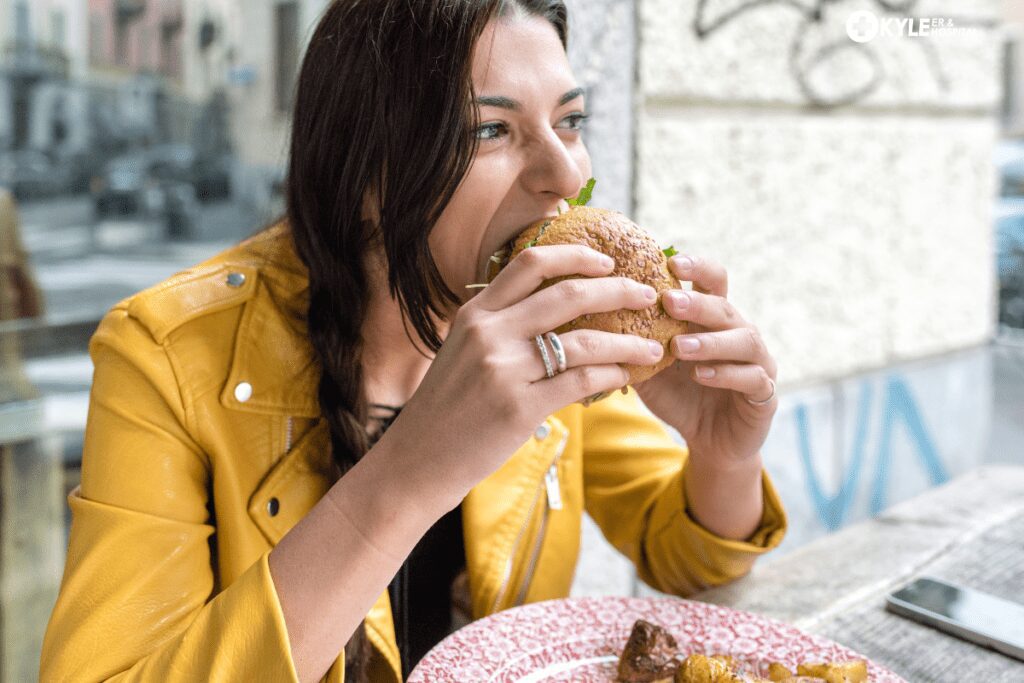 A woman in a yellow jacket bites into a large sandwich outdoors.