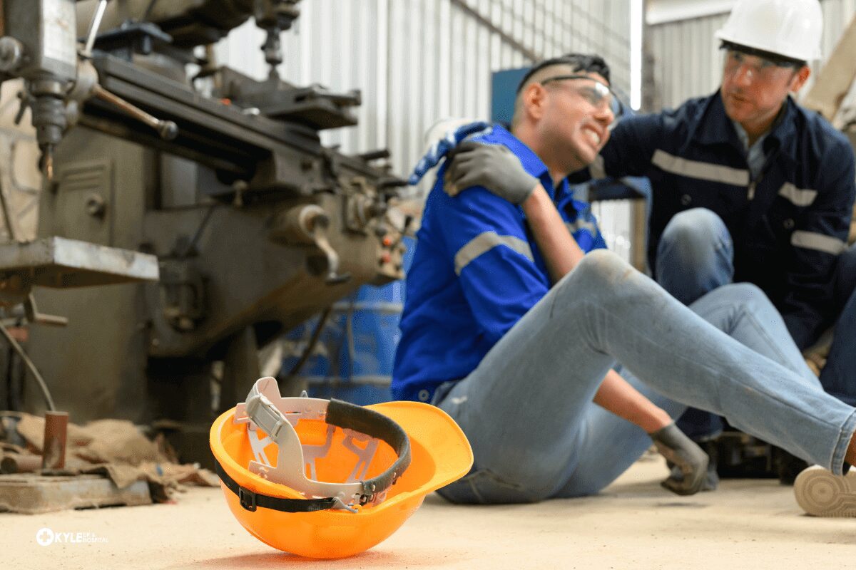Injured worker sitting on the floor near machinery with a hard hat on the ground.
